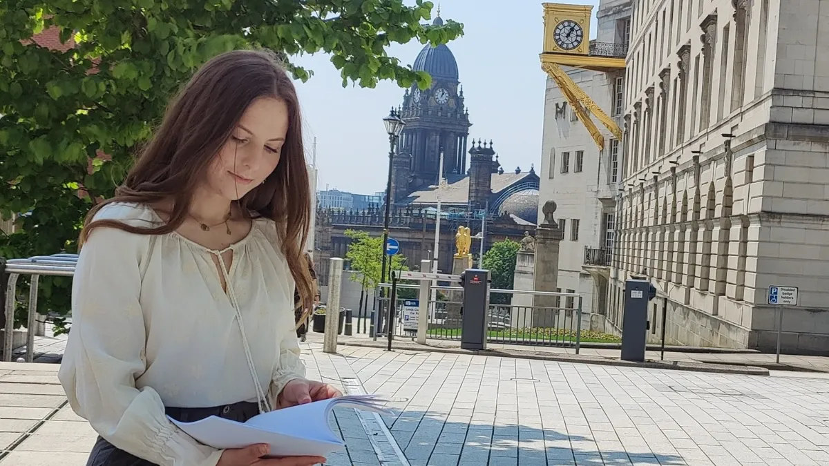 A woman in a white blouse sitting down and looking at a document on a sunny day, with the Leeds Town Hall behind her..