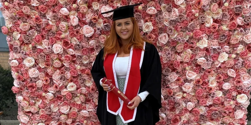 Girl with red hair in a graduation gown stands with scroll against flower drop background.