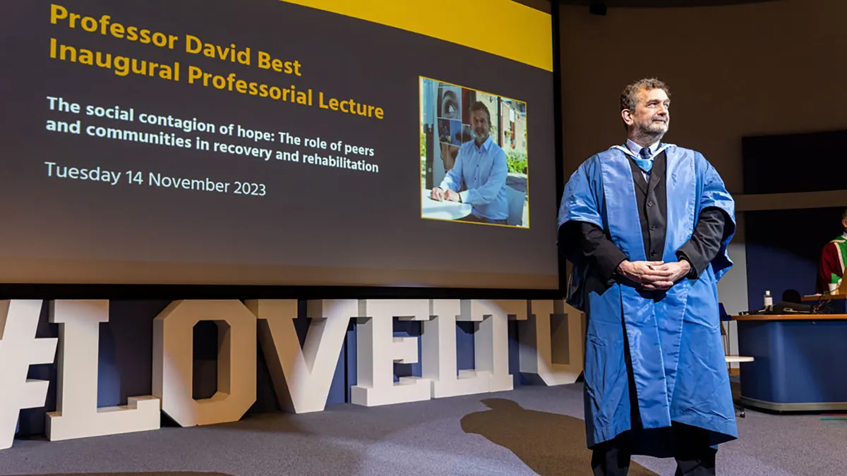 An academic in a blue gown standing in front of a screen with the title Professor David Best Inaugural Professorial Lecture written in yellow letters..