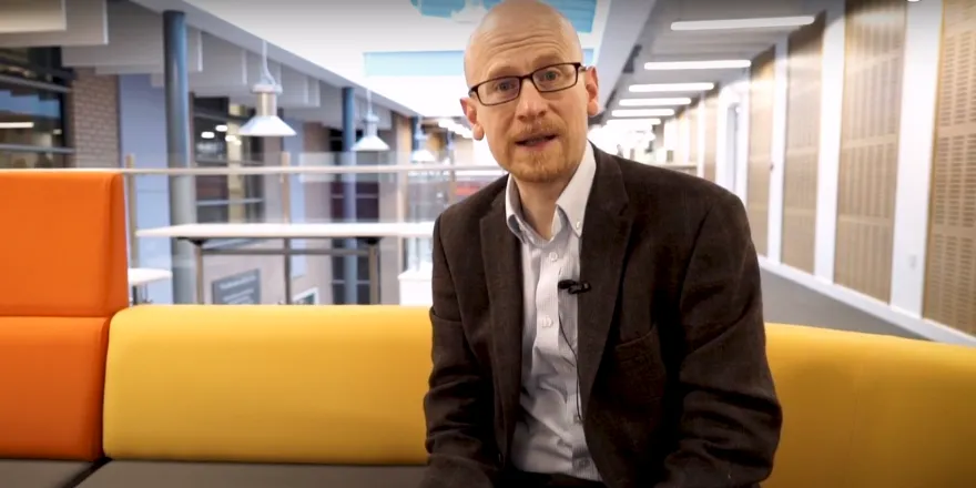 Male wearing glasses, blazer and shirt sits on yellow sofa.