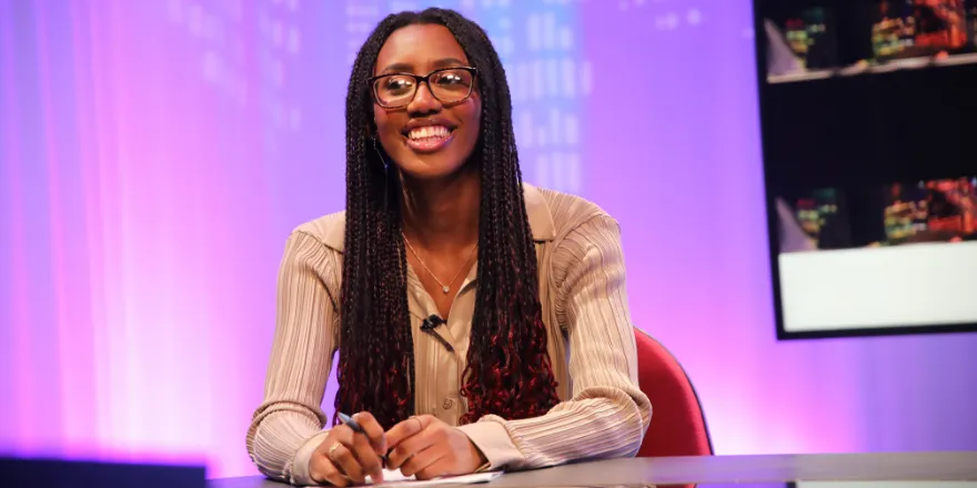 Female student sits at news desk smiling.