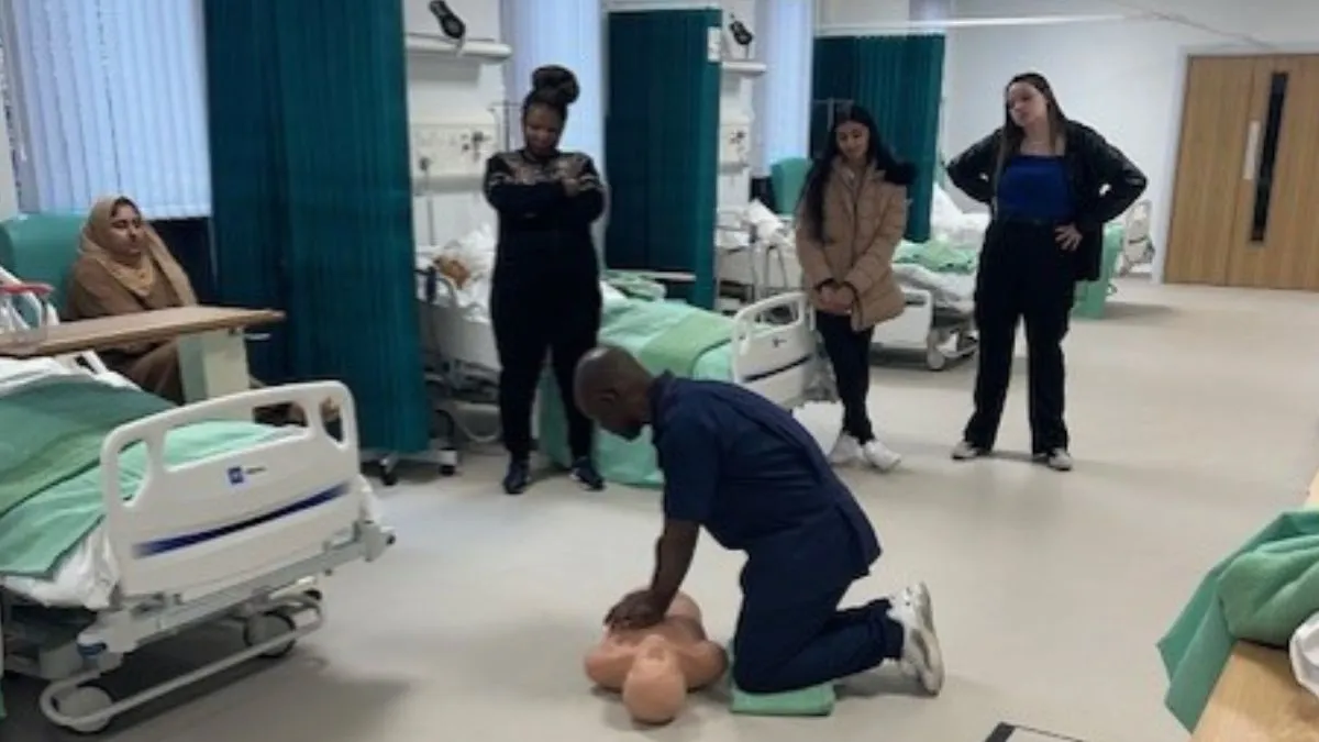 A man on his knees demonstrates chest compressions on a dummy as four students watch.