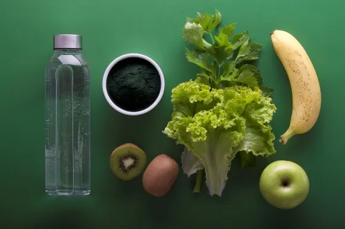Water bottle, coffee, kiwi, egg, green fruit and vegetables laid out on green background.