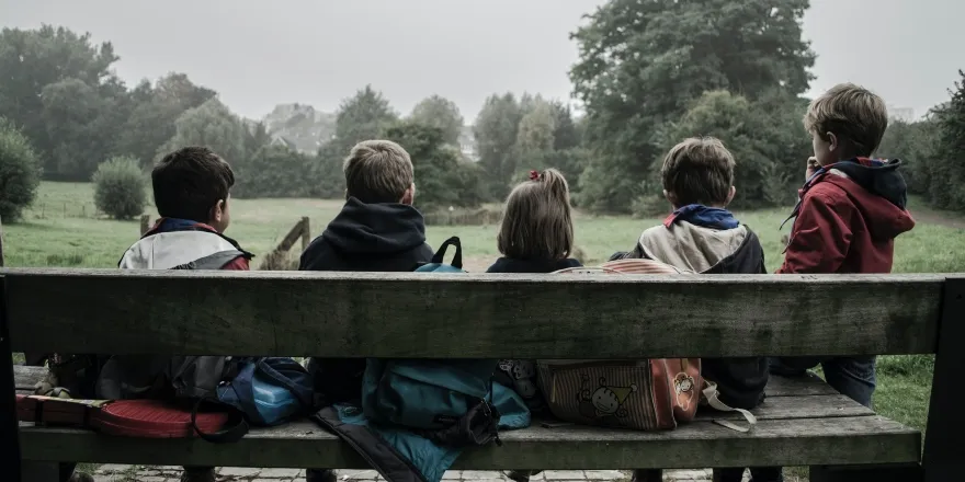 Five children sit on a wooden bench looking at green trees.