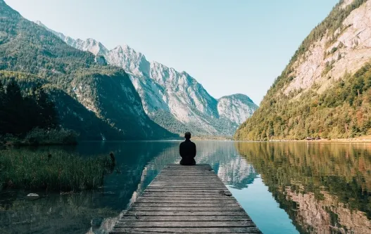 Man sitting at the edge of a dock with mountains .