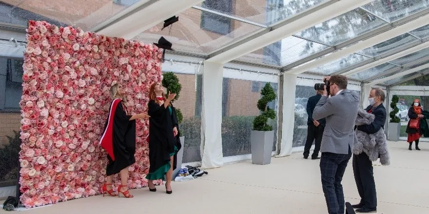 Graduates celebrating in black gowns and caps in front of pink flower wall.