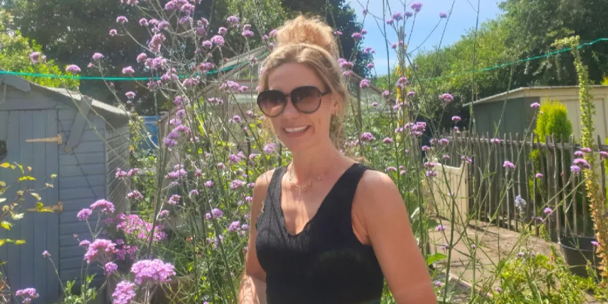 Female wearing black vest top stands in front of flowers with sunglasses on in sunshine.