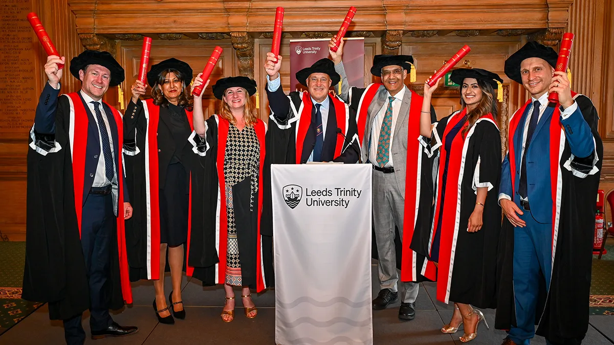 A group of people in graduation caps and gowns, holding up graduation scrolls behind a lectern covered with a white Leeds Trinity University branded cloth..