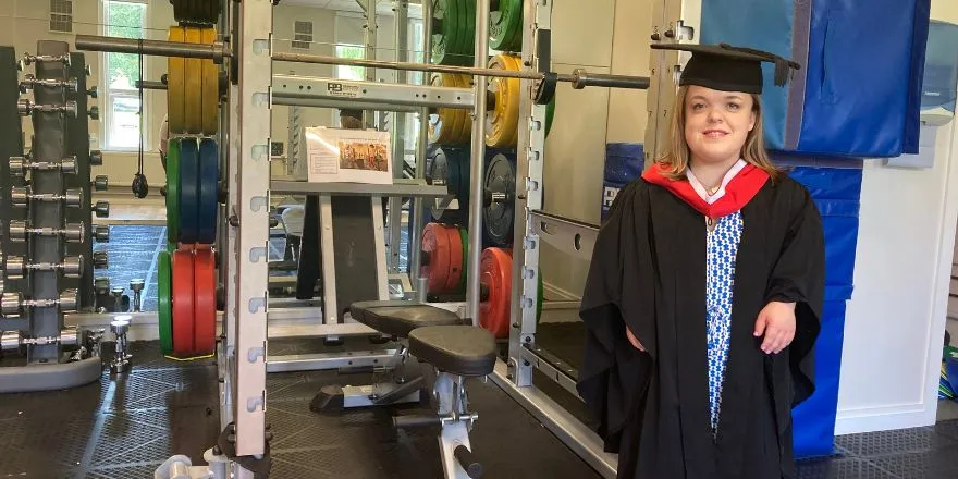 A student wearing a graduation cap and gown stands in a gym.