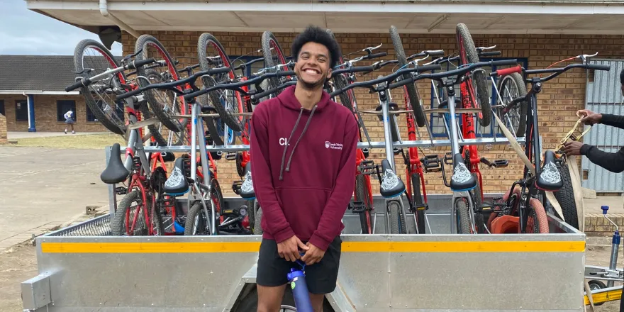 A male student wearing a burgundy hoodie smiles in front a bike rack.