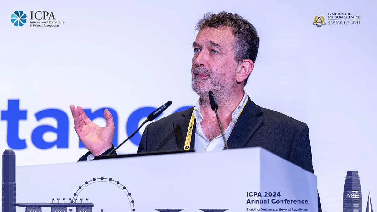 A male academic giving a speech at a conference in Singapore, behind a white lectern and wearing a black blazer..