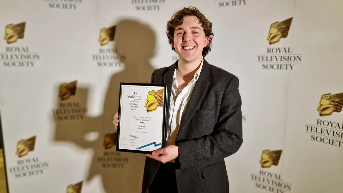 A student in a black suit smiling and holding an award in front of a Royal Television Society backdrop..