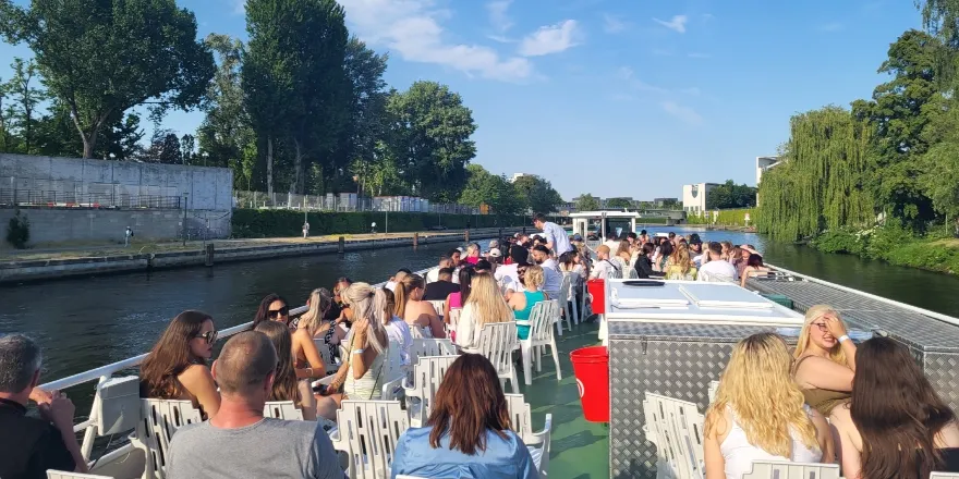 A boat tour in Berlin, with people sitting on a green deck. The river is lined with trees and the sky is blue on a sunny day..