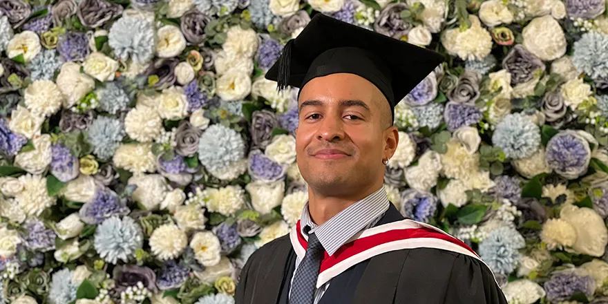 A mixed-race young man in graduation cap and gown posing in front of a wall covered in blue and white flowers..