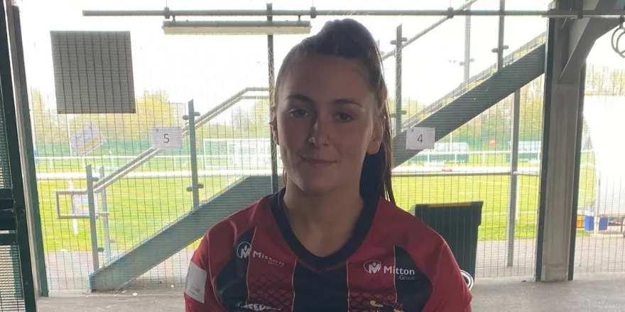 Female with brown hair tied back stands in rugby shirt in front of window.