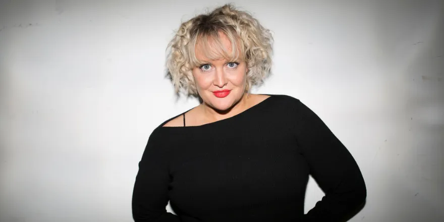 Woman with blonde curly short hair stands against white background wearing black off the shoulder long sleeved top.