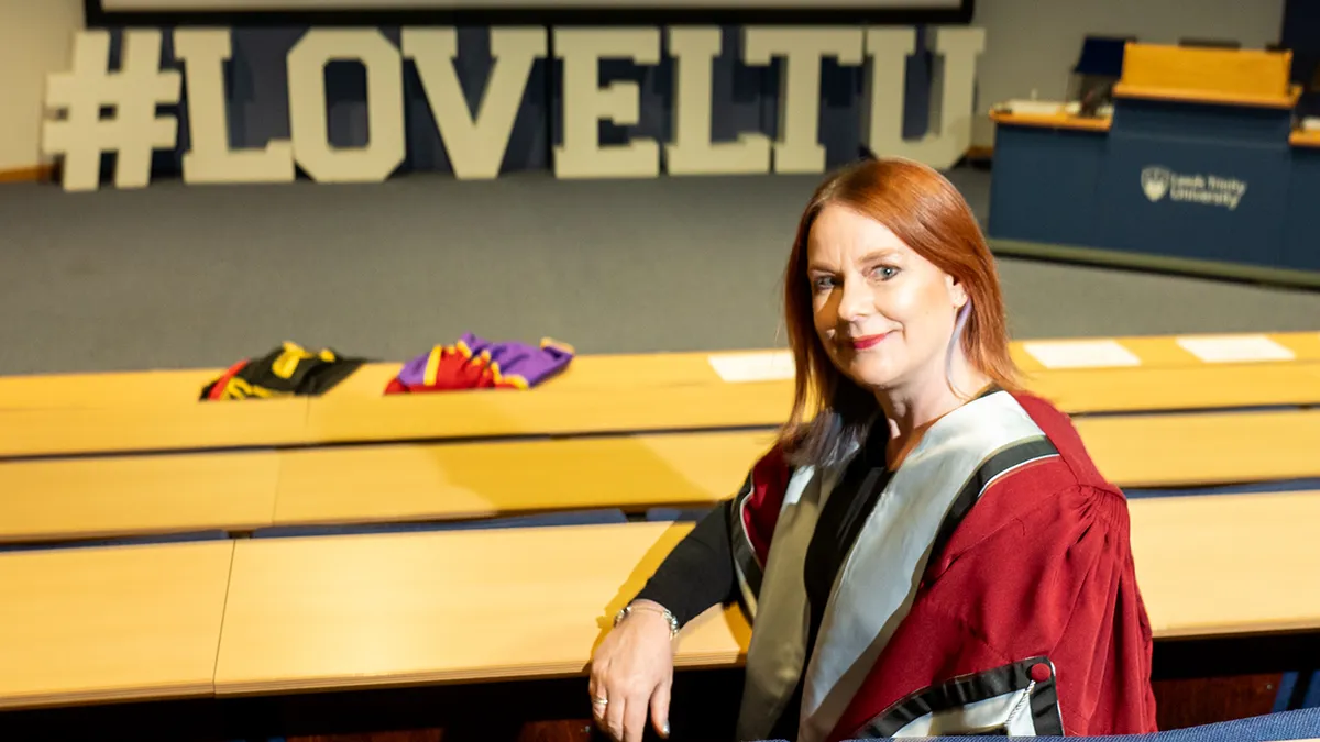 An academic in a red gown sitting in a lecture theatre in front of a sign that reads Love LTU..