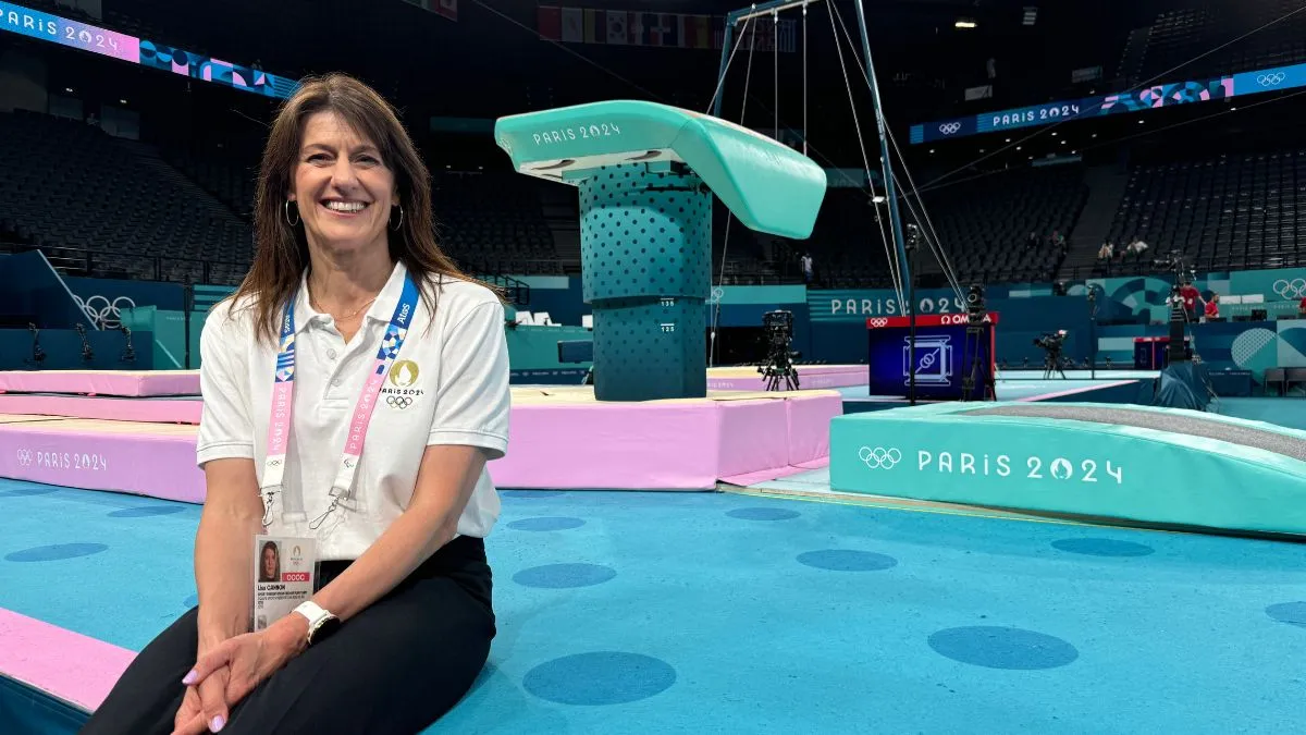 A woman with dark hair and a white top sits on a blue floor in front of gymnastics apparatus.