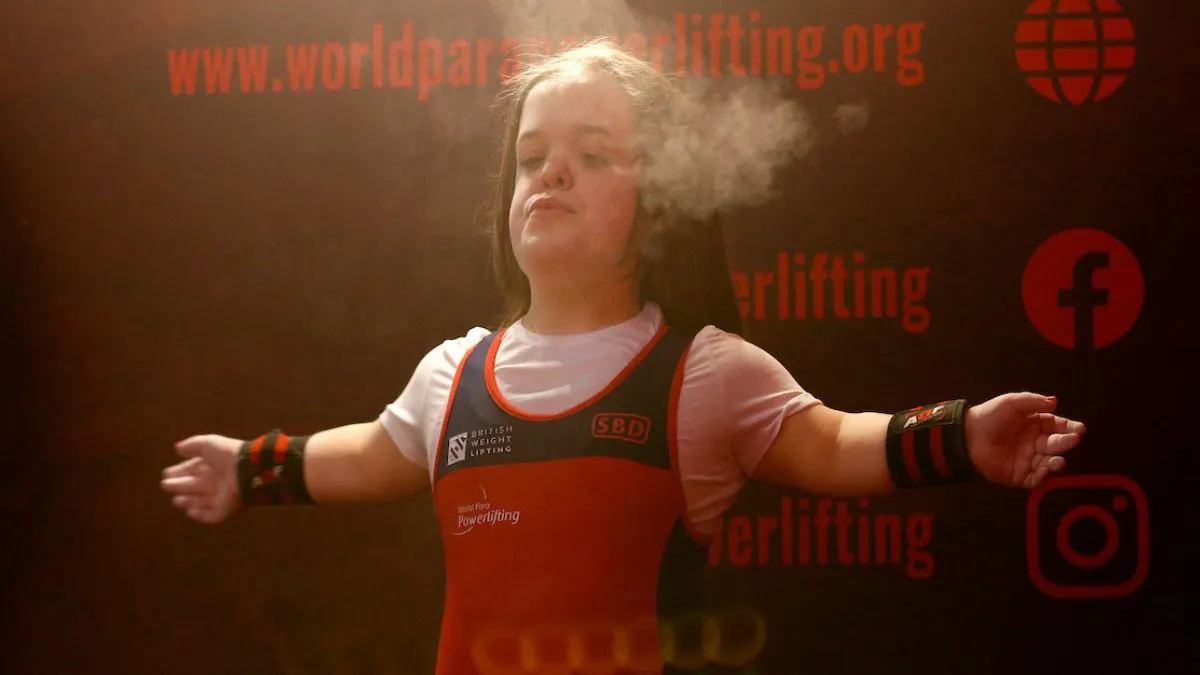 A woman with dark hair stands with her arms outstretched in front of a dark wall.