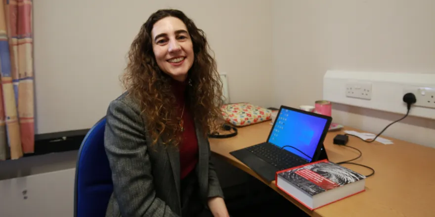 Female with dark curly hair sits at table with laptop and book.