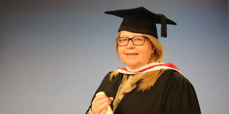 Female mature student with glasses poses for picture with graduation cap and gown, holding scroll.