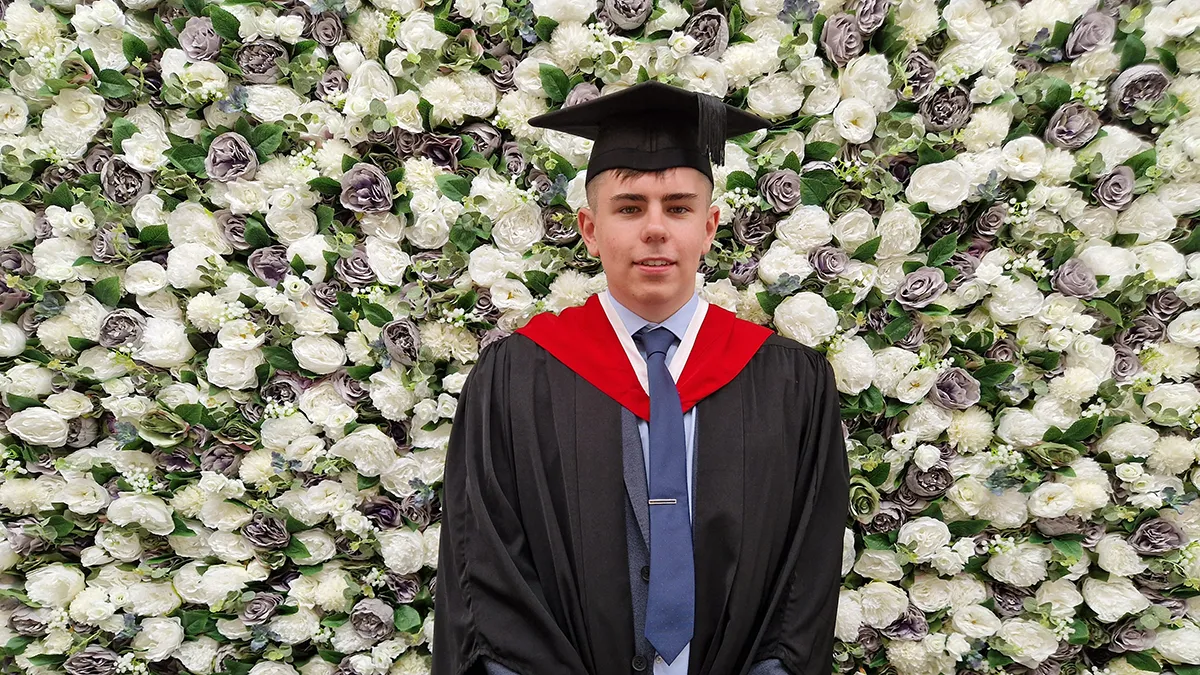 A student in graduation cap and gown smiling in front of a flower wall..