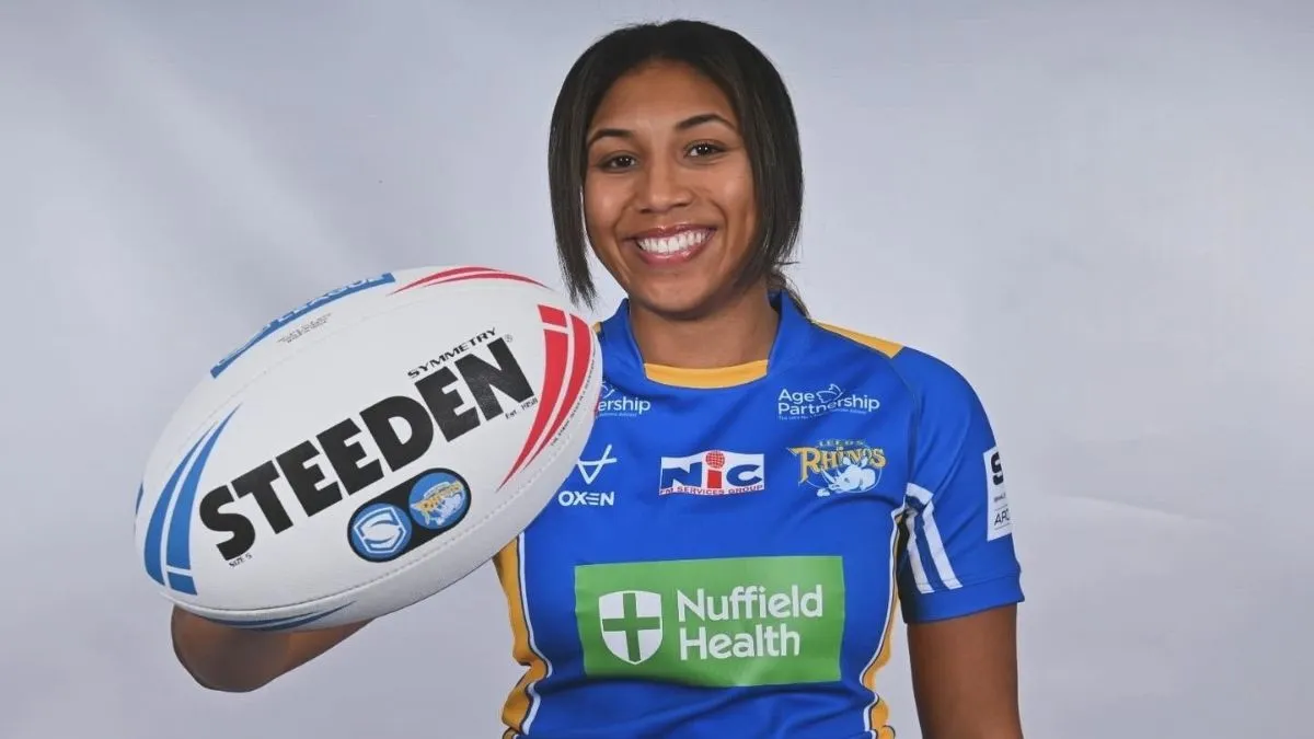 A woman with dark hair wearing a blue top smiles and holds out a rugby ball in front of her.