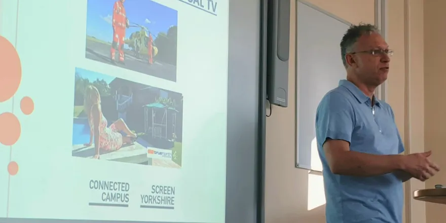 Man with grey hair and sky blue t-shirt stands presenting in front of screen.