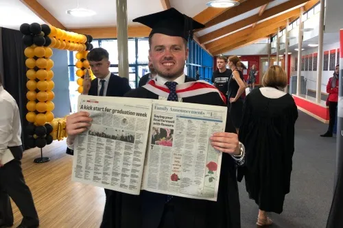 Tom pictured in cap and gown holding up newspaper.