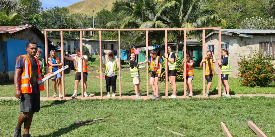 Ten volunteers wearing orange and green high-vis jackets pose for a picture as they build a wooden frame..