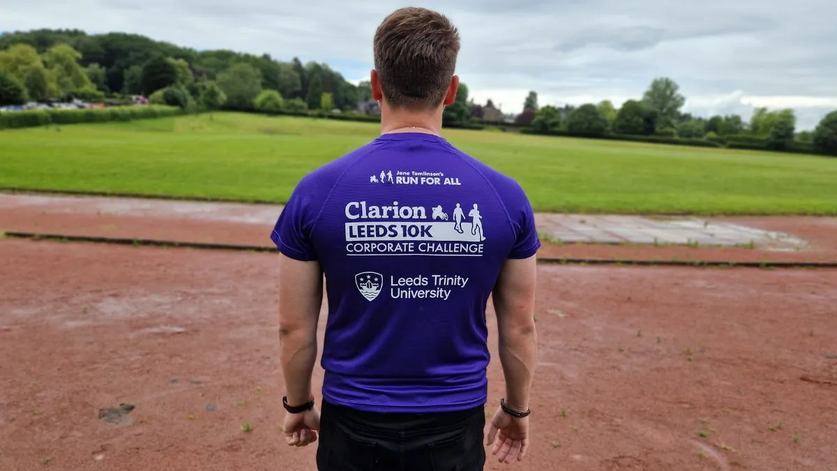 A man with dark hair wearing a purple tee shirt stands on a running track.