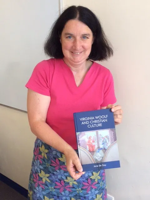 Professor Jane de Gay, with shoulder length dark hair in a pink top. Holding her book Virginia Woolf and Christian Culture..